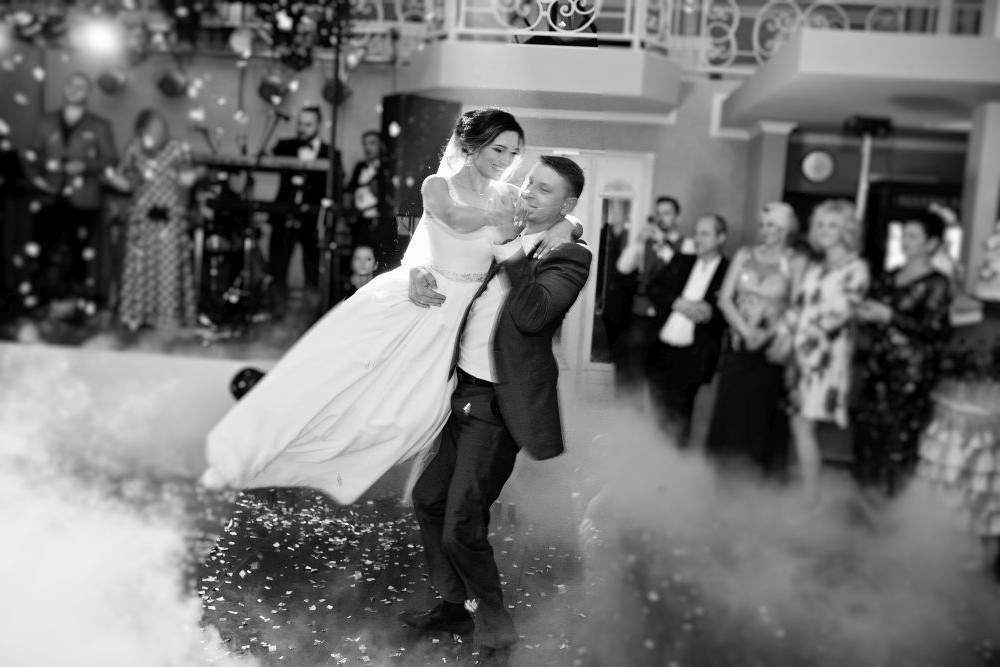 Im warm beleuchteten Ballsaal hebt der Bräutigam während des Hochzeitstanzes die Braut tanzend hoch. Der Boden ist mit Konfetti bestreut, während die Gäste voller Ehrfurcht zuschauen. Dieser in Schwarzweiß festgehaltene freudige Moment wirkt zeitlos.