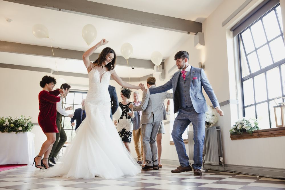 Eine Braut in einem weißen Kleid tanzt fröhlich den Hochzeitstanz mit einem Mann in einem grauen Anzug auf einem karierten Boden. Im Hintergrund tanzen mehrere Gäste in Abendgarderobe. Luftballons schweben über dem Boden und Blumen schmücken den Raum und verstärken die Atmosphäre einer Hochzeitsfeier.