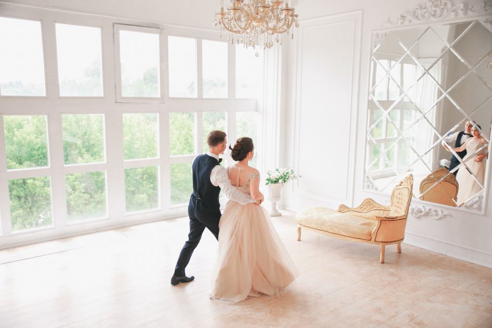 Der Hochzeitstanz findet in einem hellen Raum mit großen Fenstern und einem Kronleuchter statt. Das Spiegelbild von Braut und Bräutigam schimmert in einem großen Spiegel, während draußen üppige grüne Bäume schwanken. Ein verzierter Stuhl steht elegant neben dem Spiegel und trägt zur bezaubernden Atmosphäre bei.