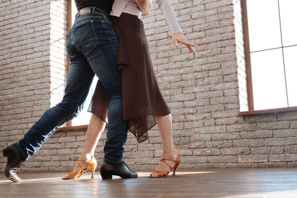 Zwei Personen tanzen eng aneinander gekuschelt in einem rustikalen Raum mit großen Fenstern und perfektionieren ihre Bewegungen für den Hochzeitstanz. Eine trägt Jeans und schwarze Schuhe, die andere einen braunen Rock mit hellbraunen Absätzen. Sonnenlicht fällt durch die Fenster und beleuchtet den Holzboden.