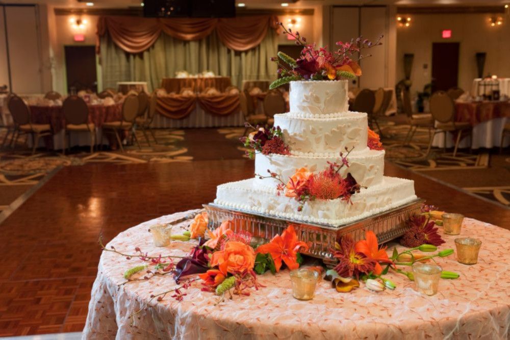 Eine dreistöckige weiße Hochzeitstorte, dekoriert mit orangefarbenen und dunkelroten Blumen, steht auf einem runden Tisch mit einer gemusterten Tischdecke. Im Hintergrund ist ein Ballsaal mit Tischen und Stühlen, gedämpftem Licht und weichen Vorhängen zu sehen.