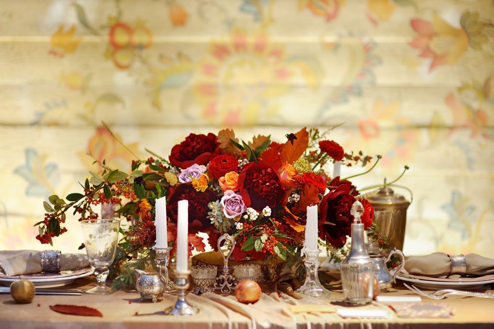 Ein dekorativ gedeckter Tisch mit roten, orangefarbenen und rosa Blumenarrangements in der Mitte. Auf dem Tisch stehen zwei weiße Kerzen, umgeben von Silberbesteck, einer Teekanne und Obst. Im Hintergrund ist eine Wand mit Blumenmuster zu sehen.