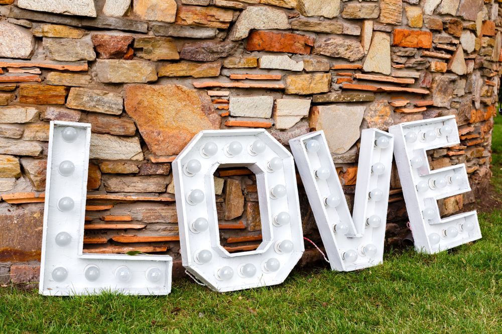 Große weiße Buchstaben mit der Aufschrift „LOVE“ sind auf dem Gras vor einer bunten Steinmauer zu sehen. Die Buchstaben sind auf dem Boden angeordnet, wobei jeweils runde Glühbirnen ihre Form umreißen.