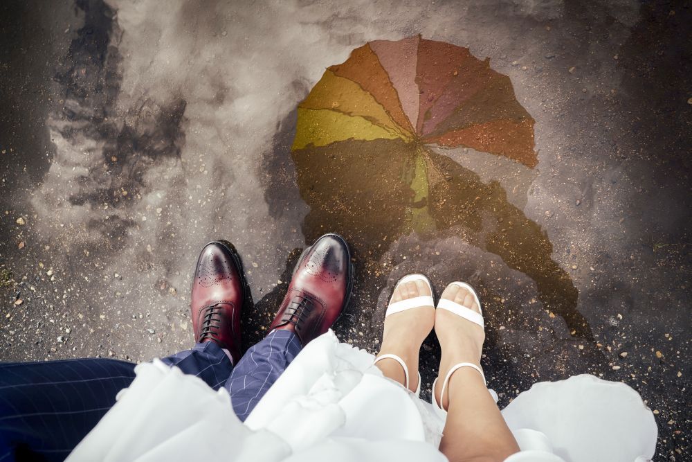 Ein Paar polierte Abendschuhe und weiße Sandalen mit Absatz stehen nebeneinander auf dem nassen Bürgersteig. Ihr Spiegelbild erscheint neben einem bunten Regenschirm in einer Pfütze und bildet einen lebhaften Kontrast zum dunklen Boden.
