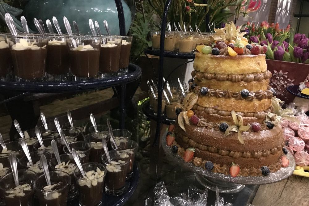 Eine Dessertauslage zeigt eine dreistöckige Torte, dekoriert mit Beeren und Fruchtscheiben, neben mehreren Gläsern Schokoladenmousse, garniert mit Nüssen, jeweils mit einem Löffel. Im Hintergrund sind Blumen und weitere Desserts zu sehen.
