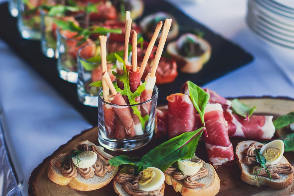 Es wird eine Auswahl an Vorspeisen angeboten, darunter in Prosciutto eingewickelte Grissini in einem Glas, mit Sahne und halbgekochten Wachteleiern belegte Baguettescheiben sowie verschiedene mit Gemüse garnierte Bruschetta-Sorten auf einer Holzplatte.