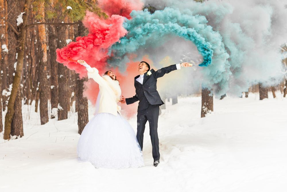 Eine Braut im weißen Kleid und ein Bräutigam im schwarzen Anzug stehen in einem verschneiten Wald. Sie halten Rauchbomben in den Händen, die Braut mit rotem Rauch und der Bräutigam mit grün-blauem Rauch, die bunte Schwaden in die Winterlandschaft zaubern.