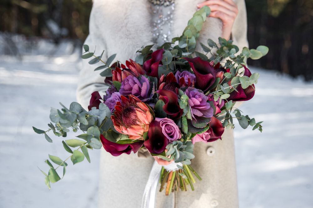 Eine Person in einem hellen Mantel hält vor einem schneebedeckten Hintergrund einen Strauß gemischter Blumen, darunter dunkelrote und violette Blüten mit Eukalyptusblättern.