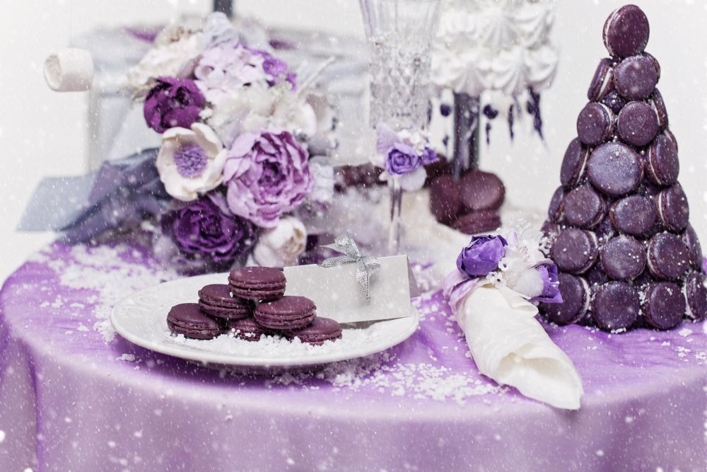 Auf einem mit violetten Akzenten dekorierten Tisch stehen ein Stapel violetter Macarons, die zu einem Kegel angeordnet sind, ein Teller mit Macarons, ein Strauß rosa und violetter Blumen und eine in Silber verpackte Geschenkkarte. Die Tischdecke ist violett und mit Schneeflocken verziert.
