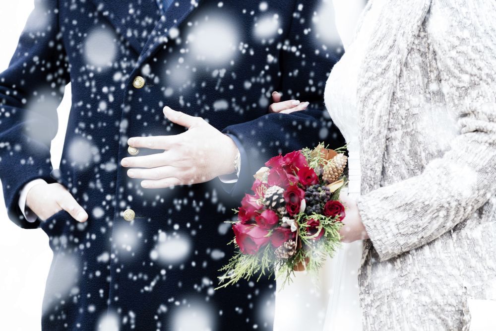 Ein Paar steht zusammen im fallenden Schnee. Die Person links trägt einen dunklen Mantel mit goldenen Knöpfen und ihre Hand ruht auf dem Arm der anderen Person. Die Person rechts hält einen Strauß roter Blumen und Grünpflanzen und trägt ein strukturiertes weißes Outfit.