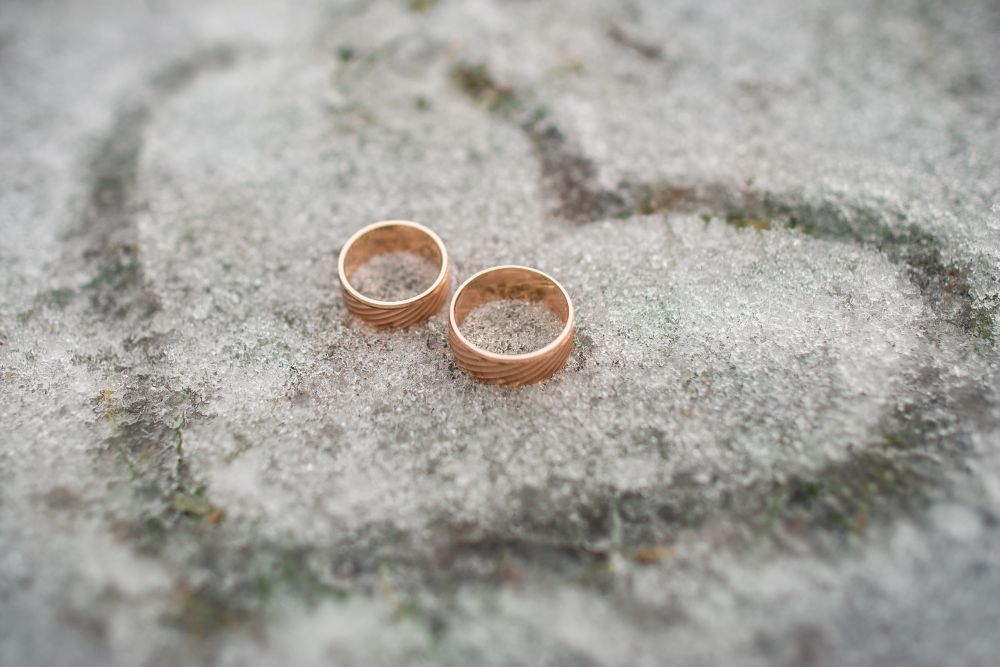 Auf einer frostigen Oberfläche liegen zwei goldene Ringe. In das Eis darunter ist schwach die Form eines Herzens eingezeichnet.