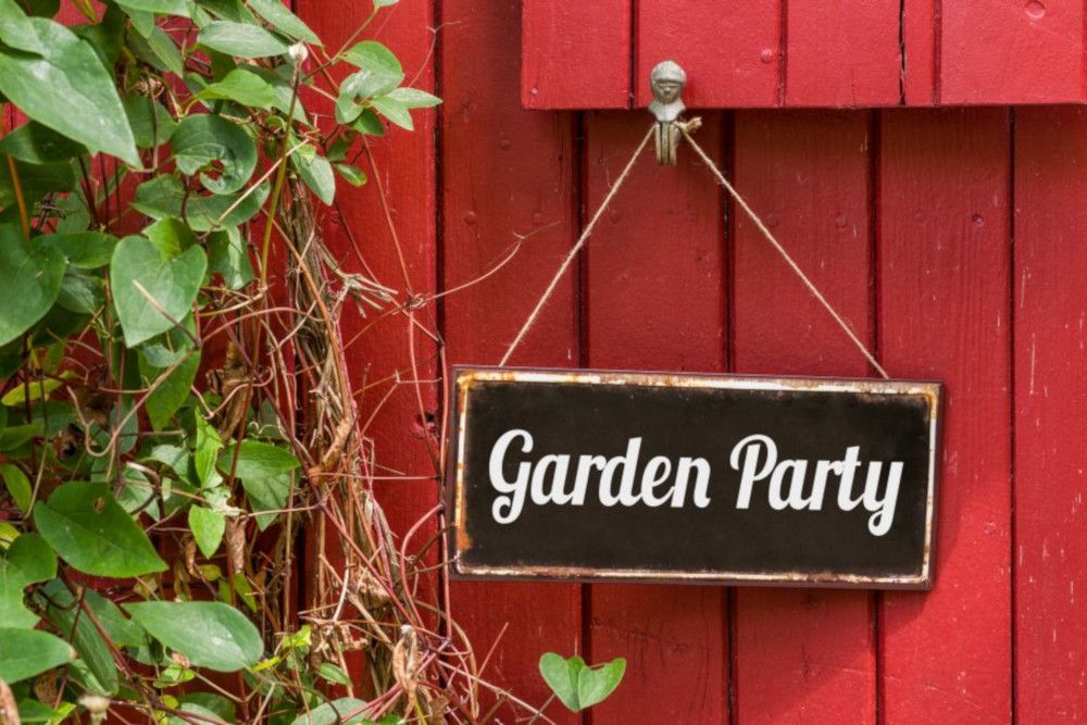 Ein Holzschild mit der Aufschrift „Gartenparty“ hängt an einer roten Wand und schafft eine einladende Atmosphäre, die an „Heiraten im Frühling“ erinnert. Grüne Blätter und Ranken wachsen in der Nähe und verleihen der Szene eine natürliche Note.