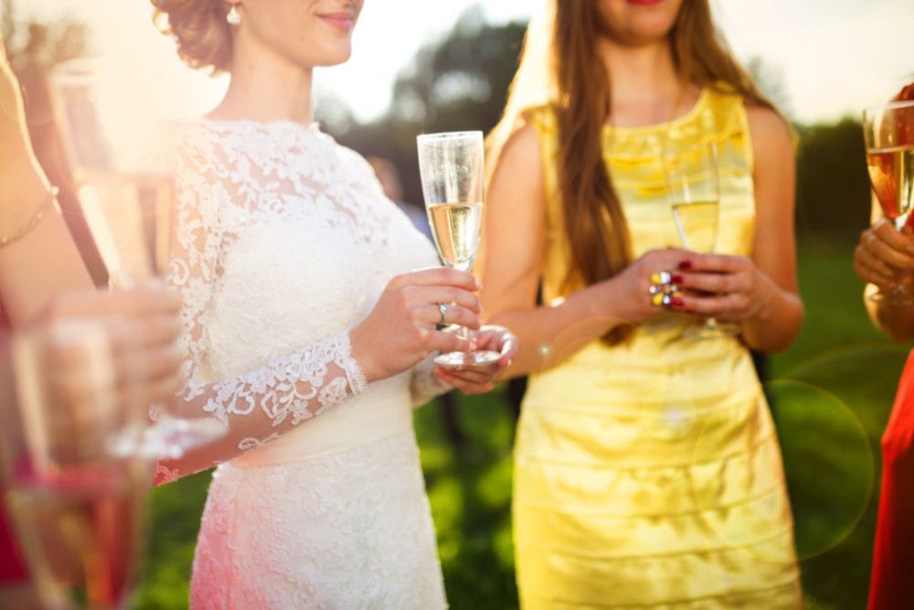 Eine Gruppe von Frauen in eleganten Kleidern stößt im Freien mit Champagnergläsern an und feiert „Heirate im Frühling“. Die Frau im Vordergrund trägt ein langärmeliges Spitzenhochzeitskleid, das die Romantik der Jahreszeit verkörpert. Im Hintergrund ist eine sonnenbeschienene Rasenfläche zu sehen.