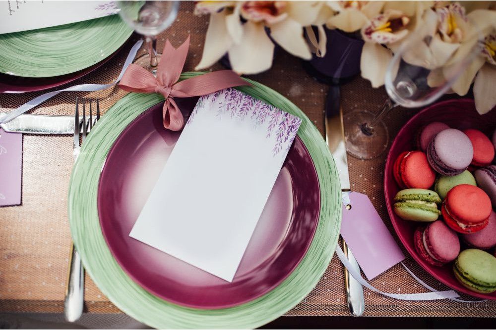 Ein gedeckter Tisch mit einem lila-grünen Teller, darauf eine mit Blumen gestaltete Speisekarte, die mit einer rosa Schleife zusammengebunden ist – perfekt für alle, die im Frühling heiraten möchten. Daneben bereichern eine Schale mit bunten Macarons und ein Blumenarrangement die Szene. Besteck und ein Weinglas stehen auf der strukturierten Tischdecke.
