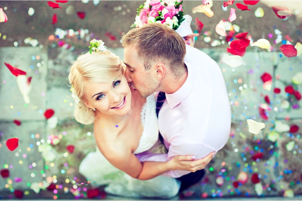 Ein glückliches Paar umarmt sich an seinem Hochzeitstag, umgeben von bunten Blütenblättern, die auf dem Boden verstreut sind, und fängt die Essenz von „Heiraten im Frühling“ ein. Die Braut lächelt in die Kamera, während der Bräutigam sie sanft auf die Wange küsst. Beide strahlen in ihrem Hochzeitskleid vor Glück.