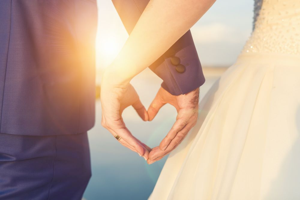 Braut und Bräutigam stehen eng beieinander und formen mit ihren Händen ein Herz, während die Sonne untergeht und einen warmen Schein über ihre Frühlingshochzeit wirft. Das Kleid der Braut ist mit kleinen, schimmernden Details verziert, perfekt für „heiraten im Frühling“.