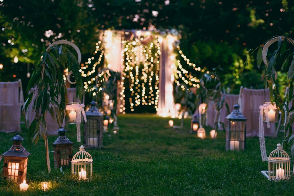 Eine romantische Hochzeitszeremonie im Freien bei Nacht, perfekt für „Heiraten im Frühling“. Der Mittelgang ist mit hängenden Laternen, Bändern und kleinen weißen Käfigen geschmückt. Lichterketten hängen elegant über einem Blumenbogen und verbreiten ein warmes, einladendes Licht.