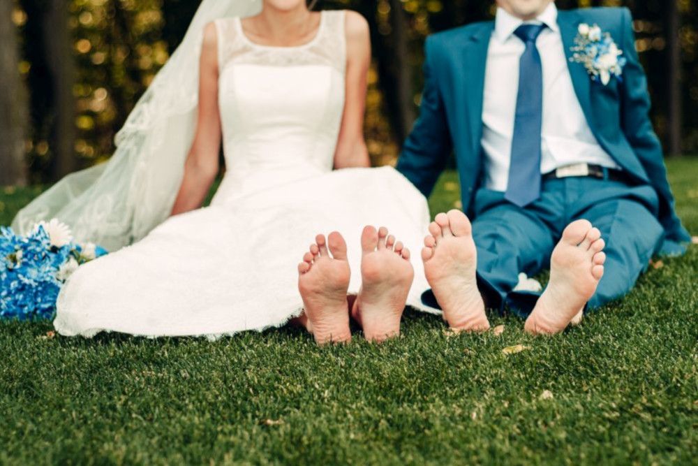 Braut und Bräutigam sitzen barfuß auf dem Gras und verkörpern damit die Essenz von „Heiraten im Frühling“. Die Braut trägt ein weißes Kleid und einen Schleier, während der Bräutigam einen blauen Anzug mit Krawatte trägt. Neben ihnen liegt ein Strauß blauer und weißer Blumen, der ihr ruhiges Frühlingsbild vervollständigt.