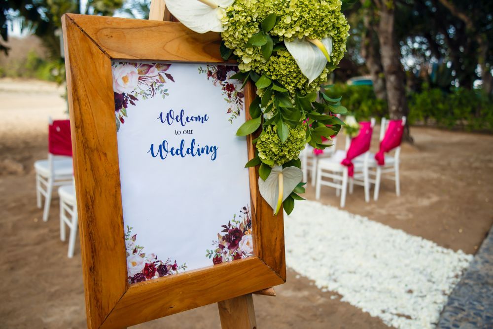 Auf einer hölzernen Staffelei hängt ein mit Blumen geschmücktes Schild mit der Aufschrift „Willkommen zu unserer Hochzeit“. Im Hintergrund sind weiße Stühle mit rosafarbenen Bezügen und ein mit weißen Blütenblättern gesäumter Gang zu sehen, die draußen zwischen üppigen Bäumen stehen – perfekt für „Heirate im Sommer“.