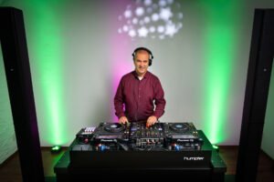 DJ Slick, Hochzeits- und Event-DJ, steht in einem burgunderroten Hemd und mit Kopfhörern hinter einem professionellen DJ-Pult und bedient die Regler, während grüne und violette Lichter für eine ausgelassene Party-Atmosphäre sorgen.