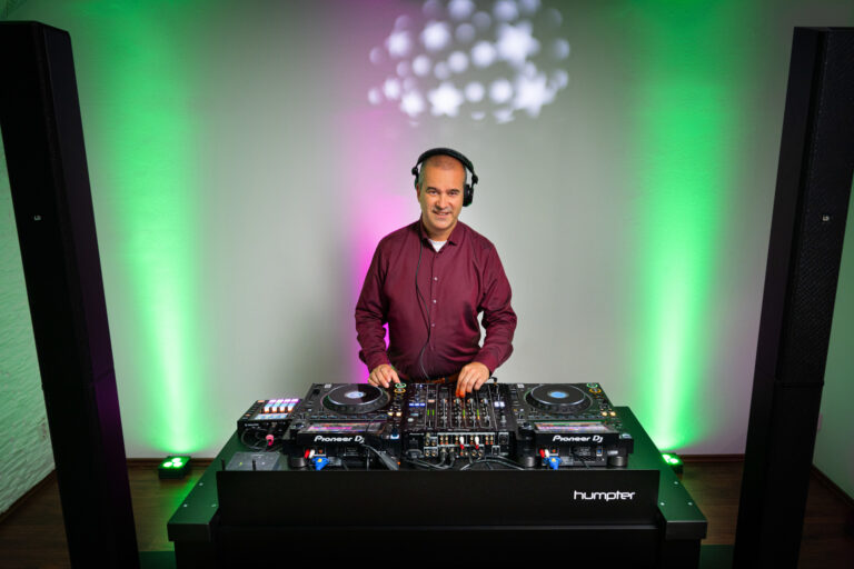 DJ Slick, Hochzeits- und Event-DJ, steht in einem burgunderroten Hemd und mit Kopfhörern hinter einem professionellen DJ-Pult und bedient die Regler, während grüne und violette Lichter für eine ausgelassene Party-Atmosphäre sorgen.