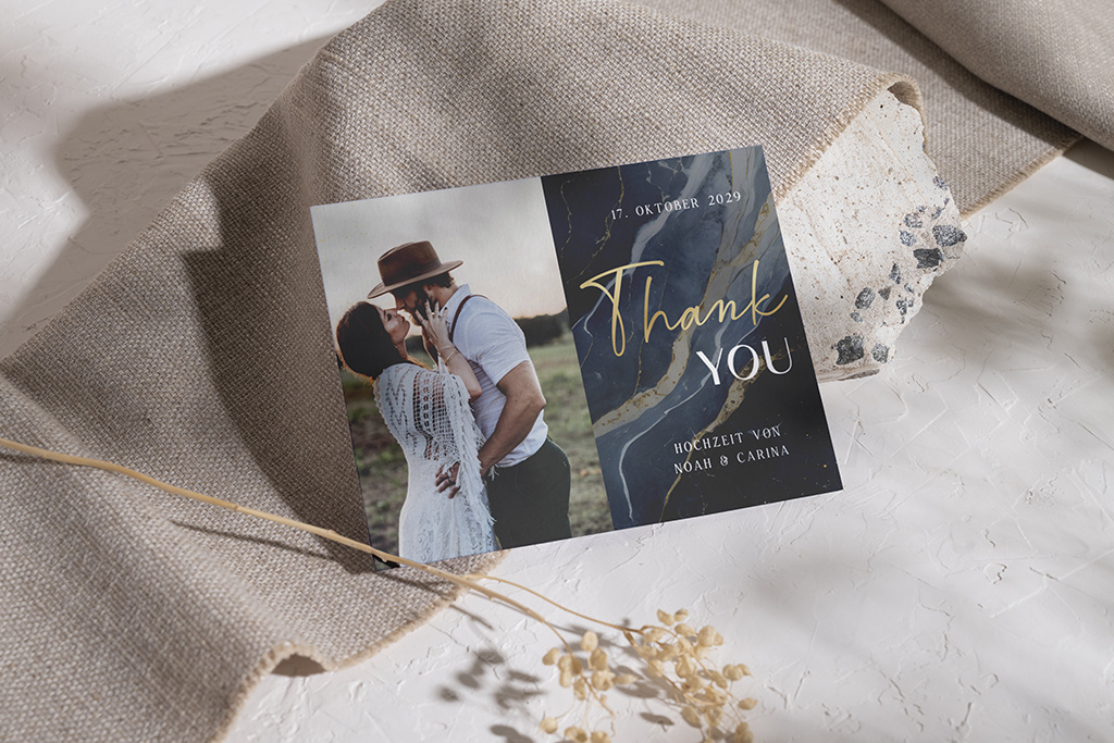 Eine Dankeskarte zur Hochzeit mit einem sich umarmenden Paar auf der linken Seite und dem Text „Danke“ auf der rechten Seite in eleganter Handschrift. Sie enthält das Datum „17. Oktober 2029“ und die Namen „Noah & Carina“. Sie ist auf einem strukturierten Stoff platziert.