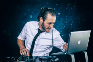 Ein DJ mit Kopfhörern benutzt einen Laptop und Mischpultausrüstung. Er trägt ein weißes Hemd und eine schwarze Krawatte. Der Hintergrund ist dunkel mit subtilen Lichteffekten.