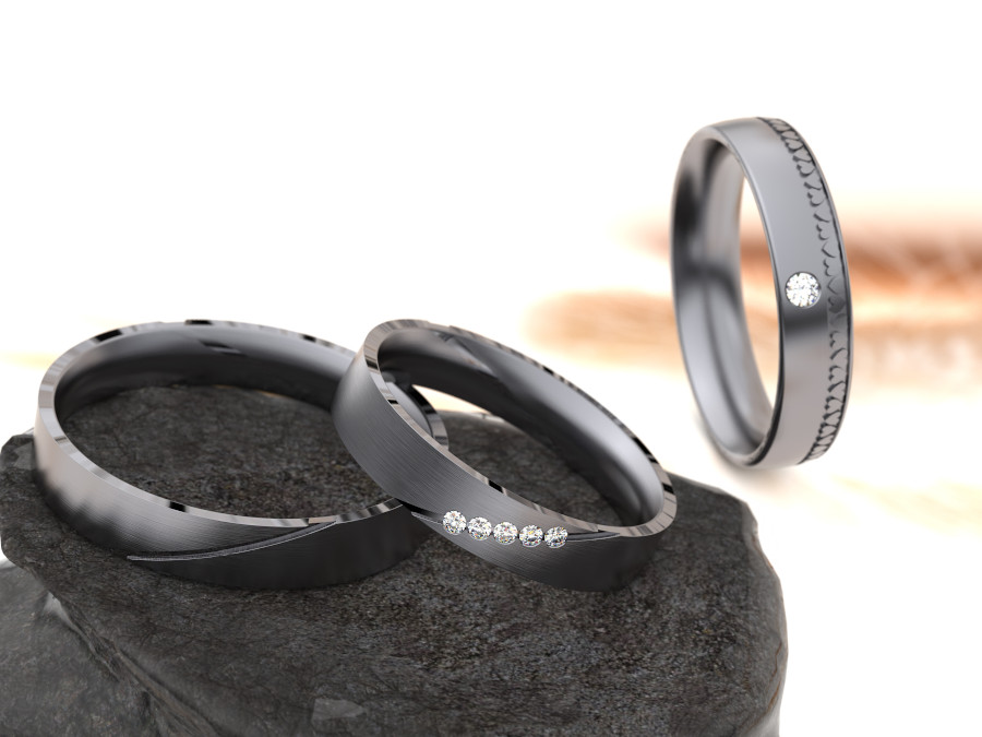 Drei elegante Ringe aus Tantal vor einem unscharfen Hintergrund. Zwei Ringe liegen auf einer Steinoberfläche und sind mit kleinen Edelsteinen verziert, während der dritte Ring aufrecht steht und einen einzelnen Edelstein aufweist.