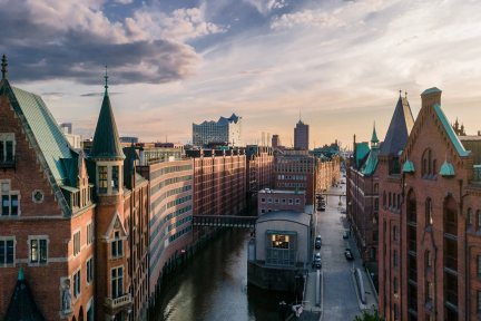 Luftaufnahme des Hamburger Speicherstadtviertels. Die Szene zeigt historische Lagerhäuser aus rotem Backstein, einen dazwischen verlaufenden Kanal und in der Ferne die moderne Elbphilharmonie unter einem teilweise bewölkten Himmel.