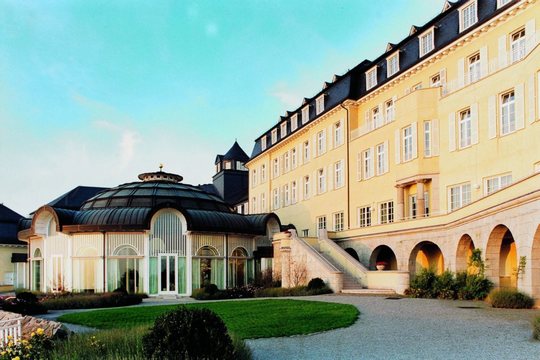 Ein großes, elegantes Hotel mit klassischem architektonischem Design verfügt über einen großen Eingang, gewölbte Fenster und eine kuppelförmige Struktur auf der linken Seite. Das Gebäude ist von einem gepflegten Rasen und Wegen unter einem klaren Himmel umgeben.
