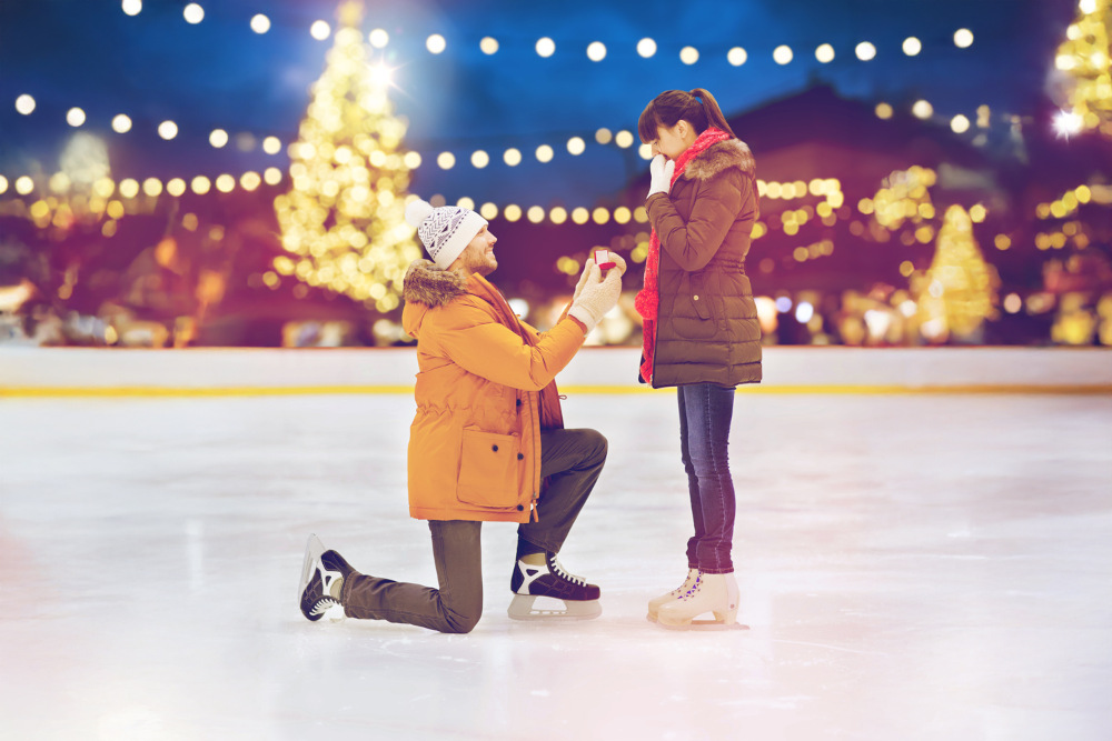 Heiratsantrag zu Weihnachten. Ein Mann kniet auf einer Eisbahn und hält eine Ringschachtel in der Hand, um einer Frau einen Heiratsantrag zu machen. Beide tragen Winterkleidung. Im Hintergrund sind Lichterketten und beleuchtete Bäume zu sehen.