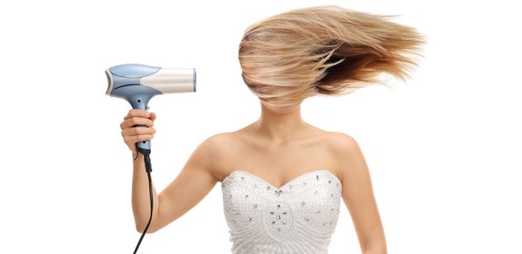 Eine Frau in einem trägerlosen Kleid hält einen Haartrockner. Ihr blondes Haar ist in ihr Gesicht geweht und verdeckt es. Der Hintergrund ist schlicht weiß.