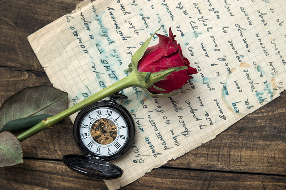 Eine rote Rose und eine geöffnete Taschenuhr liegen auf einem Stück altem Papier mit handgeschriebenem Text. Die Uhr zeigt römische Ziffern und zeigt ihr inneres Zahnrad. Die Gegenstände liegen auf einer Holzoberfläche.