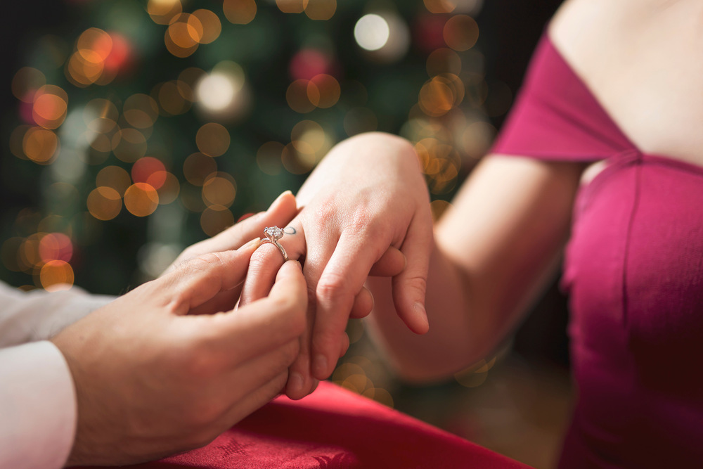 In einer Nahaufnahme steckt eine Person einer anderen Person einen Ring an den Finger. Das Paar sitzt an einem Tisch mit einer roten Tischdecke, bei sanfter Beleuchtung, mit einem verschwommenen Hintergrund eines geschmückten Weihnachtsbaums mit Bokeh-Licht.
