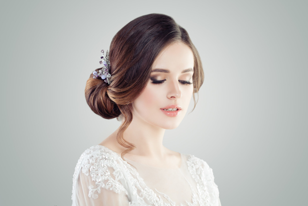 Eine Braut mit einem eleganten Haarknoten und einer Haarspange. Sie trägt ein weißes Spitzenkleid und hat vor einem hellgrauen Hintergrund die Augen geschlossen.