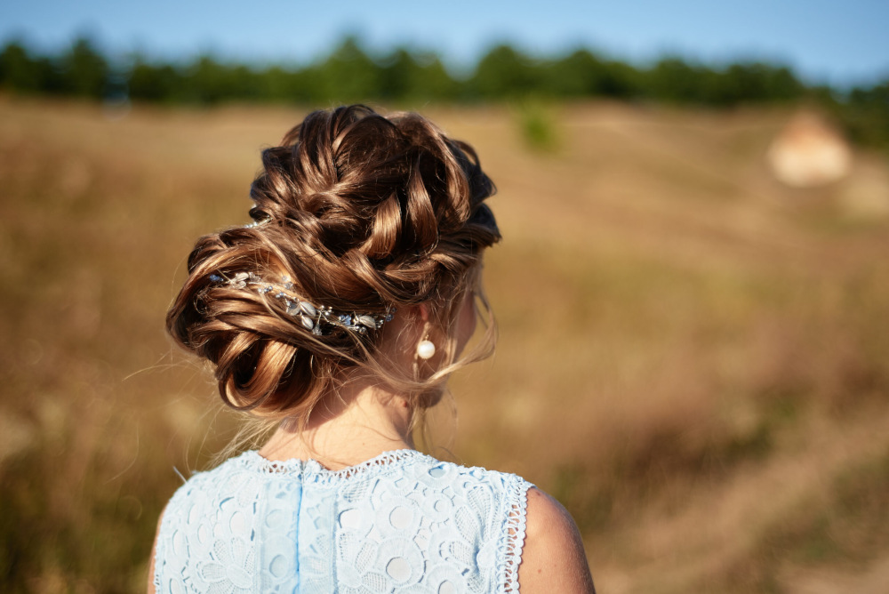 Eine Braut mit einer aufwendig geflochtenen Hochsteckfrisur, geschmückt mit einem dekorativen Haar-Accessoire, steht im Freien. Sie trägt ein hellblaues Spitzenkleid und Perlenohrringe. Der unscharfe Hintergrund zeigt ein Feld und Bäume.