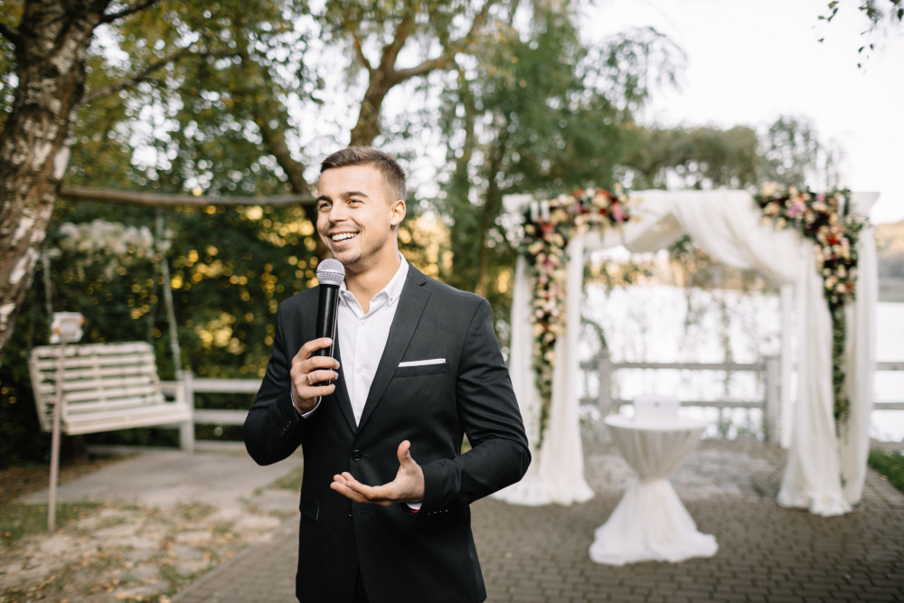 Ein Mann im Anzug hält ein Mikrofon und steht im Freien mit Bäumen im Hintergrund. Er scheint eine Rede zu halten. Hinter ihm sind ein Blumenbogen und eine Gartenschaukel zu sehen.