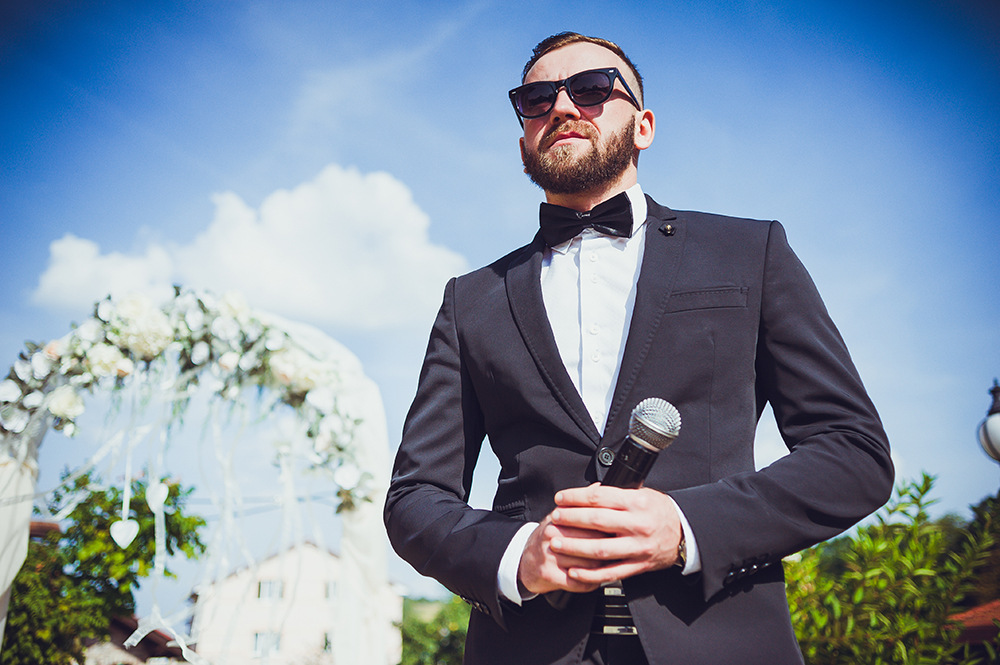 Ein Mann in schwarzem Anzug und Sonnenbrille hält draußen ein Mikrofon. Hinter ihm ist ein mit weißen Blumen und Bändern geschmückter Hochzeitsbogen. Der Himmel ist klar mit ein paar Wolken.