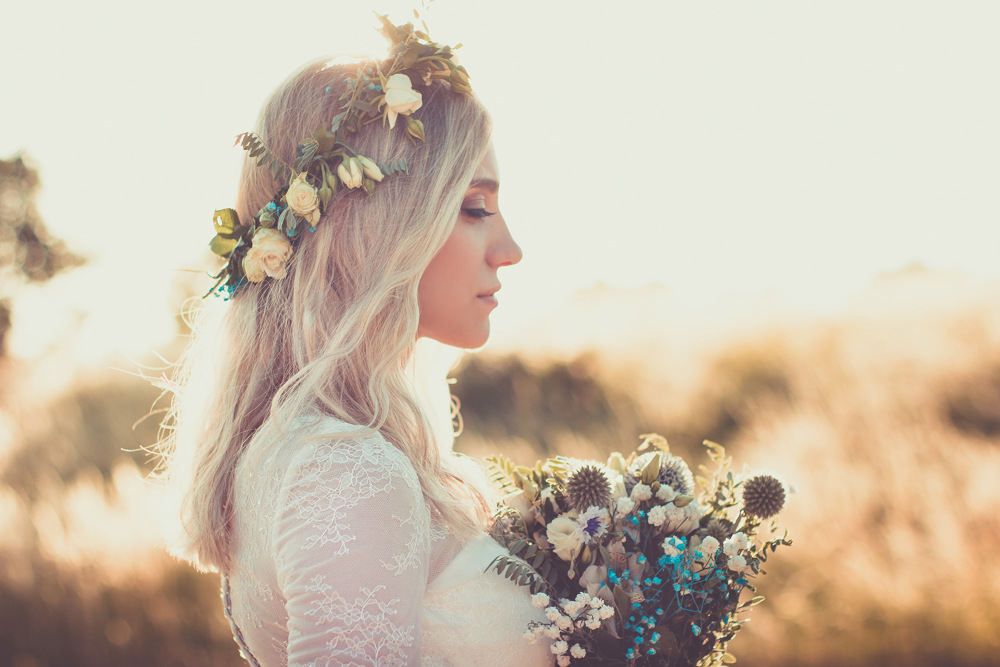 Eine Frau mit langen blonden Haaren und einem Blumenkranz steht im Profil und hält einen Blumenstrauß. Sie trägt ein Spitzenkleid und im Hintergrund ist eine verschwommene Außenkulisse mit warmem Sonnenlicht zu sehen.