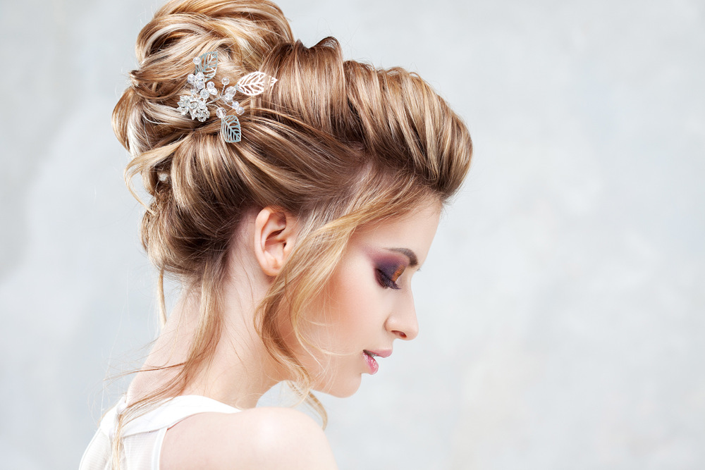 Eine Frau mit gestyltem blondem Haar in einer Hochsteckfrisur, geschmückt mit silbernen Blumenaccessoires. Sie trägt ein weißes Oberteil und rosa Lidschatten. Sie posiert vor einem weichen, hellgrauen Hintergrund und blickt nach links unten.