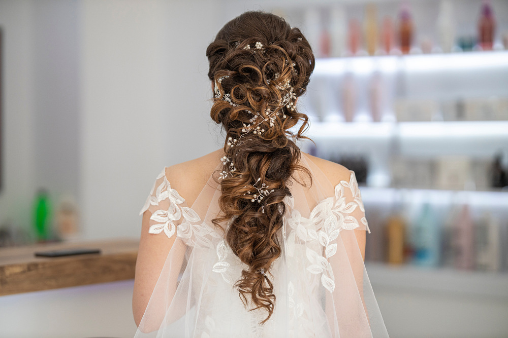 Eine Braut mit aufwendig gestyltem, langem Haar, das mit dekorativen Elementen geschmückt ist, trägt ein weißes Kleid mit Spitzendetails an den Schultern. Im Hintergrund ist ein sanft beleuchteter Raum mit Regalen zu sehen, auf denen verschiedene dekorative Flaschen ausgestellt sind.
