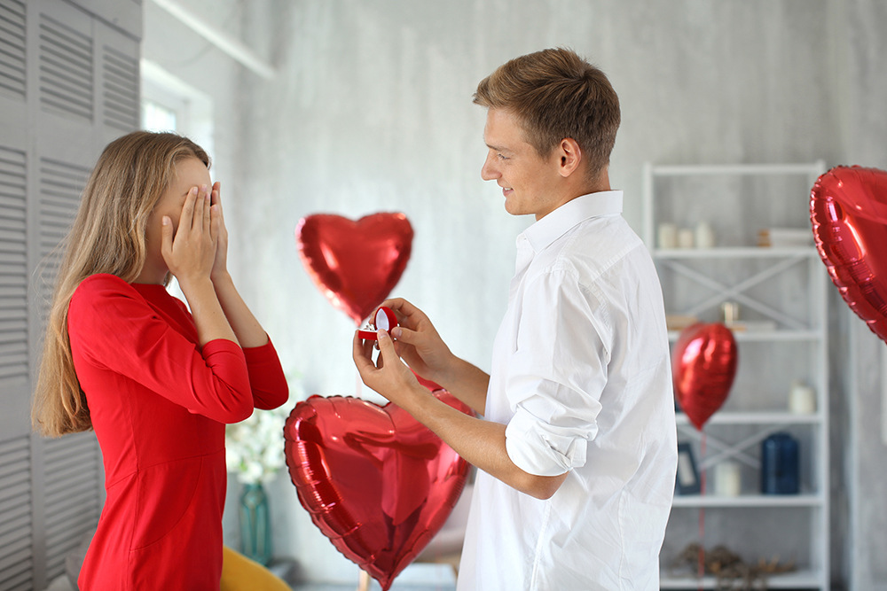Ein Mann im weißen Hemd kniet und hält einer Frau im roten Kleid, die ihr Gesicht mit den Händen bedeckt, eine geöffnete Ringschachtel hin. Rote herzförmige Luftballons schmücken den Hintergrund in einem Raum mit hellen Wänden.