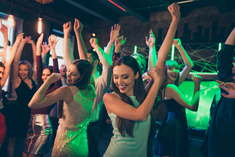 In einem schwach beleuchteten Nachtclub mit bunten Lichtern tanzen Menschen ausgelassen. Eine Frau im Vordergrund lächelt, umgeben von anderen, die die lebhafte Atmosphäre genießen.