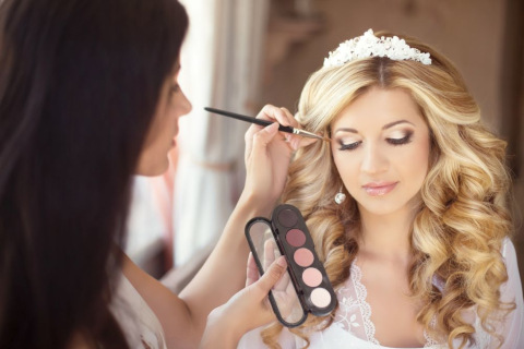 Eine Frau mit langen, lockigen blonden Haaren, einer Tiara und einem spitzenbesetzten Gewand trägt ein Make-up-Makeup-Artist und lässt sich von einer anderen Frau die Augenlider schminken. Die Visagistin hält eine Palette mit verschiedenen Lidschattentönen in der Hand.