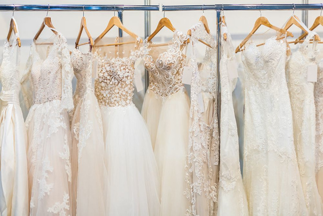 Eine Reihe eleganter Brautkleider auf hölzernen Kleiderbügeln, jedes mit aufwendigen Spitzen- und Stickereimustern. Die Kleider variieren im Stil, einige haben transparente Ärmel und Perlendetails. Sie hängen in einer gut beleuchteten Umgebung an einem metallischen Kleiderständer.