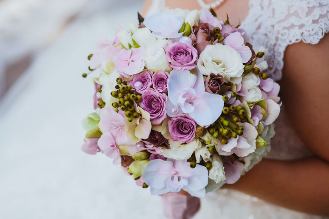Eine Nahaufnahme eines Brautstraußes mit einer Mischung aus weißen, rosa und violetten Blüten mit grünen Knospen. Die Braut trägt ein Spitzenbrautkleid.